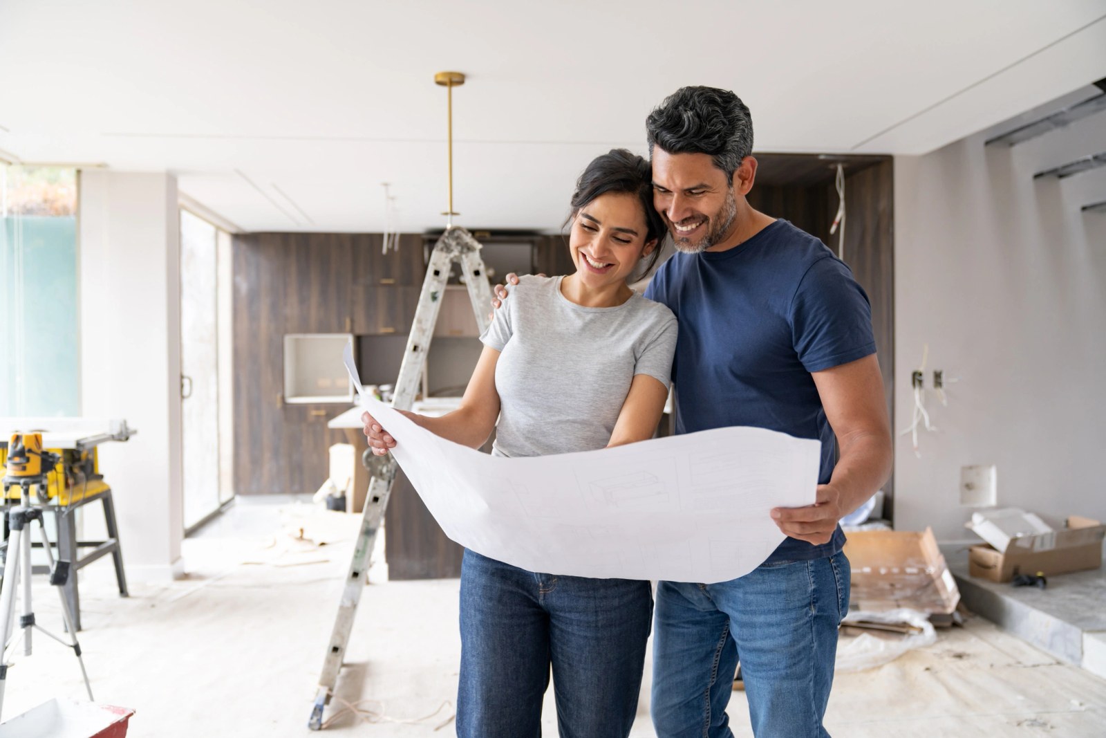 A couple reading plans in a kitchen part way through a renovation, with a stepladder and tools around them